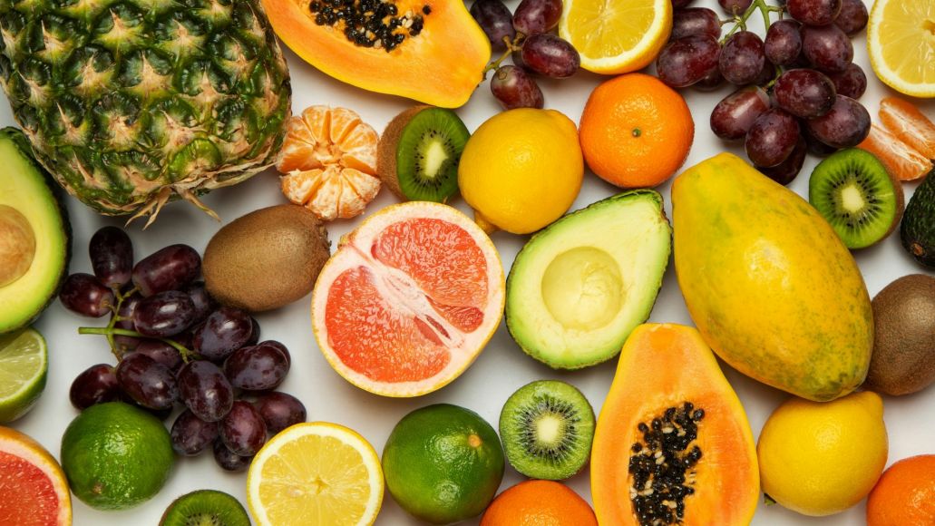 Mixed fruit on a plain surface, including avocado, kiwifruit, pineapple, grapes, limes, lemons, and others.